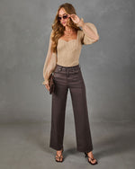 A woman modeling a beige long-sleeve top with sheer balloon sleeves and dark brown wide-leg trousers, standing against a textured gray studio backdrop.