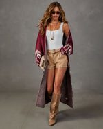 Woman wearing a long burgundy patterned cardigan, white bodysuit, tan leather shorts, and suede knee-high boots with sunglasses