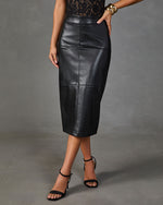 A woman shown from the back wearing a black faux-leather midi skirt with a front slit, paired with black strappy high-heeled sandals and gold bracelets, against a gray studio backdrop.