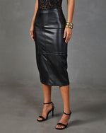 A woman shown from the back wearing a black faux-leather midi skirt with a front slit, paired with black strappy high-heeled sandals and gold bracelets, against a gray studio backdrop.