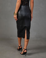 A woman shown from the back wearing a black faux-leather midi skirt with a front slit, paired with black strappy high-heeled sandals and gold bracelets, against a gray studio backdrop.