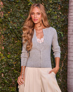 Woman in gray ribbed cardigan over lace cami, cream wide-leg trousers, holding chain-strap bag, posed outdoors against green ivy wall.