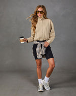 A woman modeling a beige long-sleeve zip-up jacket, black biker shorts, and a plaid shirt tied around her waist, holding a coffee cup against a gray studio background.