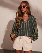 Woman wearing green striped boho blouse with balloon sleeves, cream shorts, gold sunglasses, and woven crossbody bag