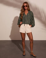 Woman wearing green striped boho blouse, cream shorts, strappy sandals, and round sunglasses, holding woven crossbody bag