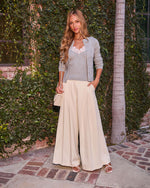 Woman in gray cardigan, lace cami, cream wide-leg trousers, white sandals, holding quilted bag outdoors
