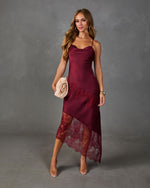 A woman models a burgundy satin slip dress with matching lace panels and sheer lace hem, styled with clear high-heeled sandals against a textured gray studio background.