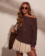 Woman wearing oversized dark brown off-shoulder knit sweater, cream pleated mini skirt with lace trim, gold accessories, and tinted sunglasses