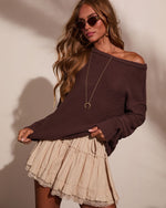 Woman wearing dark brown off-shoulder knit sweater with beige lace-trim mini skirt, gold jewelry, and oval sunglasses