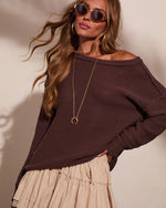 Woman wearing chocolate brown off-shoulder knit sweater with cream tiered maxi skirt, gold crescent necklace, and round sunglasses