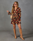 A woman modeling a long-sleeve mini dress with a red, yellow, and black floral pattern, styled with tan knee-high leather boots, a beige woven handbag, and sunglasses against a gray studio backdrop.