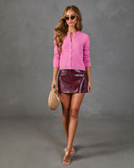 Woman wearing pink cropped cardigan, burgundy patent leather mini skirt, clear heels, holding woven clutch bag