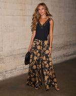 A woman stands against a textured concrete wall wearing a sleeveless black lace-trim top and wide-leg black pants with a large tan floral print. She holds a small black woven clutch.