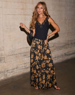 A woman posing in a black sleeveless top with floral print skirt against a concrete wall.