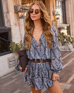 A woman wearing a blue and gray paisley mini dress with a brown belt and gold buckle, styled with sunglasses, layered necklaces, and a textured clutch, standing on a city sidewalk.