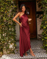 Woman in burgundy pleated one-shoulder satin maxi dress holding champagne coupe, ivy-covered entrance, gold jewelry, clear heels