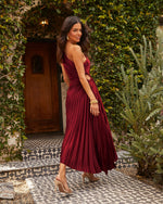 Woman in burgundy pleated midi dress with open back, gold jewelry, nude heels, posed in ivy-covered courtyard