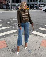 A woman stands on a city crosswalk wearing a dark olive-brown military-style jacket with gold buttons, blue jeans, and white pointed-toe heels, holding a brown clutch in an urban street setting.