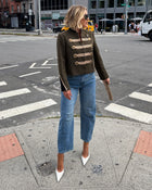 A woman stands on a city crosswalk wearing a dark olive-brown military-style jacket with gold buttons, blue jeans, and white pointed-toe heels, holding a brown clutch in an urban street setting.