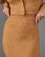 Close-up of a person wearing a fuzzy camel-colored knit cardigan and matching skirt, with both hands pulling the waistband slightly outward to show stretch. The background is a solid neutral gray.